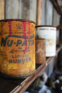 An old, rusty can of liquid rubber with vivid red paint dripping from the top stands prominently on a wooden shelf. There are other similarly aged cans visible in the background, out of focus. The setting appears to be a dusty and ambient space, possibly an old workshop or garage, with a rustic and vintage atmosphere.