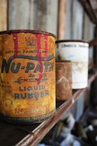 An old, rusty can of liquid rubber with vivid red paint dripping from the top stands prominently on a wooden shelf. There are other similarly aged cans visible in the background, out of focus. The setting appears to be a dusty and ambient space, possibly an old workshop or garage, with a rustic and vintage atmosphere.