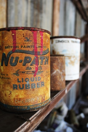 An old, rusty can of liquid rubber with vivid red paint dripping from the top stands prominently on a wooden shelf. There are other similarly aged cans visible in the background, out of focus. The setting appears to be a dusty and ambient space, possibly an old workshop or garage, with a rustic and vintage atmosphere.