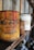 An old, rusty can of liquid rubber with vivid red paint dripping from the top stands prominently on a wooden shelf. There are other similarly aged cans visible in the background, out of focus. The setting appears to be a dusty and ambient space, possibly an old workshop or garage, with a rustic and vintage atmosphere.