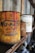 An old, rusty can of liquid rubber with vivid red paint dripping from the top stands prominently on a wooden shelf. There are other similarly aged cans visible in the background, out of focus. The setting appears to be a dusty and ambient space, possibly an old workshop or garage, with a rustic and vintage atmosphere.