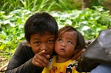 Two children are in a natural setting surrounded by lush green foliage. The older child is pointing towards something, while the younger one looks up with curiosity. They both appear to be wearing casual clothing, and the younger child is dressed in a bright yellow shirt with a cartoon motif.