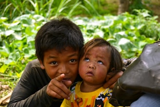 Two children are in a natural setting surrounded by lush green foliage. The older child is pointing towards something, while the younger one looks up with curiosity. They both appear to be wearing casual clothing, and the younger child is dressed in a bright yellow shirt with a cartoon motif.