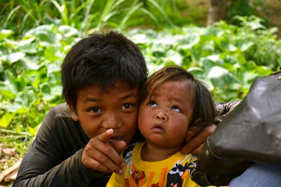Two children are in a natural setting surrounded by lush green foliage. The older child is pointing towards something, while the younger one looks up with curiosity. They both appear to be wearing casual clothing, and the younger child is dressed in a bright yellow shirt with a cartoon motif.