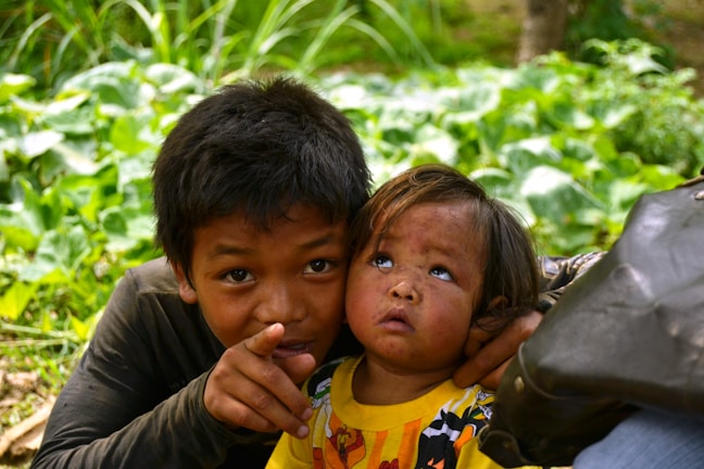 Two children are in a natural setting surrounded by lush green foliage. The older child is pointing towards something, while the younger one looks up with curiosity. They both appear to be wearing casual clothing, and the younger child is dressed in a bright yellow shirt with a cartoon motif.