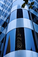 Reflective curved glass windows of a modern skyscraper exterior create an abstract design with a mirrored image of another building. The surface is smooth and sleek, with a few tree leaves visible in the upper corner.