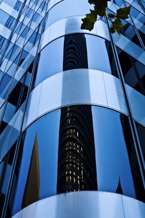 Reflective curved glass windows of a modern skyscraper exterior create an abstract design with a mirrored image of another building. The surface is smooth and sleek, with a few tree leaves visible in the upper corner.