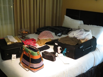 A hotel room bed is covered with an assortment of luggage and bags, including two black suitcases, a colorful striped purse, and several plastic bags. Clothes, receipts, and packaged goods are also visible on the bed. The room is dimly lit with curtains drawn behind the bed.