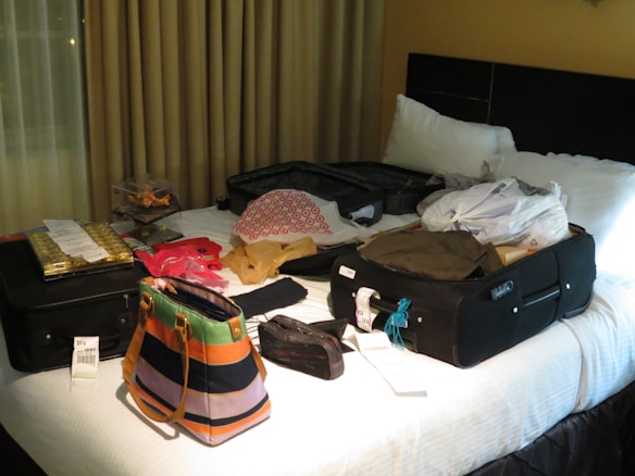 A hotel room bed is covered with an assortment of luggage and bags, including two black suitcases, a colorful striped purse, and several plastic bags. Clothes, receipts, and packaged goods are also visible on the bed. The room is dimly lit with curtains drawn behind the bed.