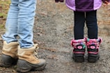 A pair of adults and children's boots stand on a gravel path; the adult boots are brown and sturdy, while the child wears colorful, patterned boots with pink accents.