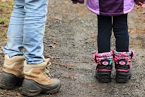 A pair of adults and children's boots stand on a gravel path; the adult boots are brown and sturdy, while the child wears colorful, patterned boots with pink accents.