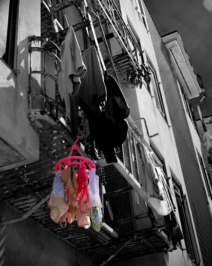 A black and white urban exterior with clothes hanging from a balcony. Brightly colored underwear, in shades of red, yellow, and blue, is prominently displayed against the monochrome backdrop. The building structure is weathered and shows multiple levels of hanging laundry.
