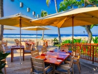 A scenic beachfront restaurant with wooden tables and chairs under large umbrellas, surrounded by lush green plants and a view of the blue ocean and white sand in the background. String lights hang between palm trees, adding to the tropical ambiance.