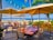A scenic beachfront restaurant with wooden tables and chairs under large umbrellas, surrounded by lush green plants and a view of the blue ocean and white sand in the background. String lights hang between palm trees, adding to the tropical ambiance.