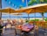 A scenic beachfront restaurant with wooden tables and chairs under large umbrellas, surrounded by lush green plants and a view of the blue ocean and white sand in the background. String lights hang between palm trees, adding to the tropical ambiance.