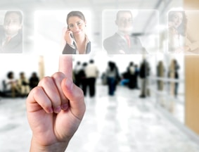 A hand is pointing to a digital interface displaying translucent images of business professionals. In the background, there are blurred figures of people standing in what appears to be a modern office or public space.