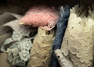 A collection of intricately detailed lace fabrics in various shades, including pink, blue, and cream. The lace is displayed in a layered manner, showcasing floral patterns and varying textures.