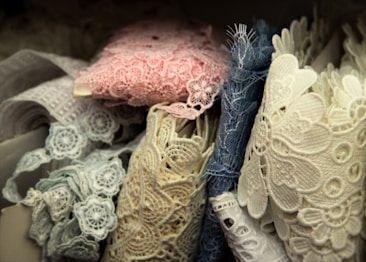 A collection of intricately detailed lace fabrics in various shades, including pink, blue, and cream. The lace is displayed in a layered manner, showcasing floral patterns and varying textures.