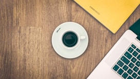 A flat-lay of a wooden table with a white coffee cup on a saucer placed near a laptop and a yellow notebook. The notebook has the word 'AWWWARDS' on it. The laptop's keyboard is partially visible in the lower right corner.
