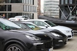 Several parked cars, including black and white models, are situated in an urban area. A black inflatable boat with branding is positioned beside the vehicles. Modern office buildings with glass windows are in the background, and a person stands near the cars.