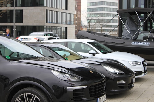 Several parked cars, including black and white models, are situated in an urban area. A black inflatable boat with branding is positioned beside the vehicles. Modern office buildings with glass windows are in the background, and a person stands near the cars.