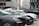 Several parked cars, including black and white models, are situated in an urban area. A black inflatable boat with branding is positioned beside the vehicles. Modern office buildings with glass windows are in the background, and a person stands near the cars.