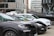 Several parked cars, including black and white models, are situated in an urban area. A black inflatable boat with branding is positioned beside the vehicles. Modern office buildings with glass windows are in the background, and a person stands near the cars.