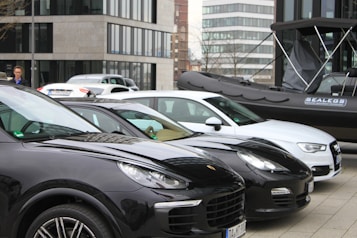 Several parked cars, including black and white models, are situated in an urban area. A black inflatable boat with branding is positioned beside the vehicles. Modern office buildings with glass windows are in the background, and a person stands near the cars.