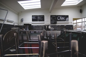 The interior of a cafe or restaurant is closed, indicated by chairs stacked upside down on tables. Black and white menu boards are displayed on the wall behind a counter. Natural light streams through the large skylights, and a window reveals parked cars outside.