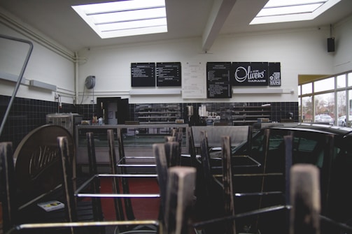 The interior of a cafe or restaurant is closed, indicated by chairs stacked upside down on tables. Black and white menu boards are displayed on the wall behind a counter. Natural light streams through the large skylights, and a window reveals parked cars outside.