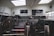 The interior of a cafe or restaurant is closed, indicated by chairs stacked upside down on tables. Black and white menu boards are displayed on the wall behind a counter. Natural light streams through the large skylights, and a window reveals parked cars outside.