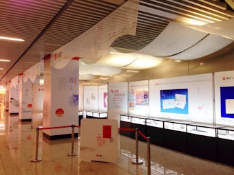 An indoor exhibition area with several display boards and glass cases, featuring information panels with text and images. The ceiling has white draped fabric, and the area is lit with overhead lights. Red ropes and stanchions are used for crowd control.