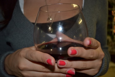 A person holds a glass of red wine with both hands. The hands have bright pink nail polish and the person is wearing a gray sweater. A necklace with a pendant resting on the chest is also visible.