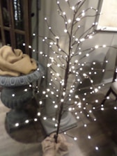 A decorative faux tree adorned with small white lights stands next to an ornate plant stand. The plant stand is draped with a tan cloth. The setting appears to be indoors with wooden flooring and relaxed ambient lighting.
