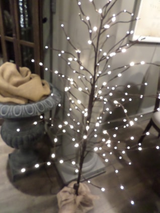 A decorative faux tree adorned with small white lights stands next to an ornate plant stand. The plant stand is draped with a tan cloth. The setting appears to be indoors with wooden flooring and relaxed ambient lighting.