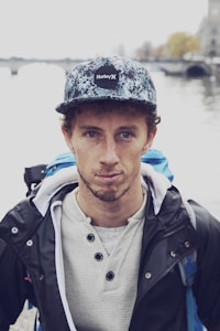 A person stands outdoors near a body of water, wearing a patterned cap with the Hurley logo, a light colored henley shirt, and a dark jacket with a blue backpack visible behind. The background features a blurred view of a river and what appears to be a bridge, with trees that have autumn-colored leaves.