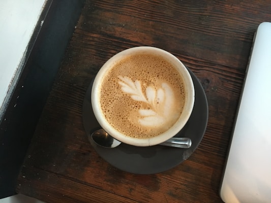 A cup of coffee with latte art in the shape of a leaf sits on a dark saucer. The cup is placed on a rustic wooden table beside a closed laptop.