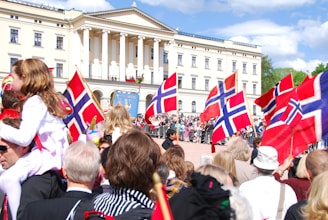 A large gathering of people is celebrating outside a grand building with columns. Many individuals are holding Norwegian flags, and a young girl is sitting on someone's shoulders. The crowd appears festive, with a diverse range of people of all ages.