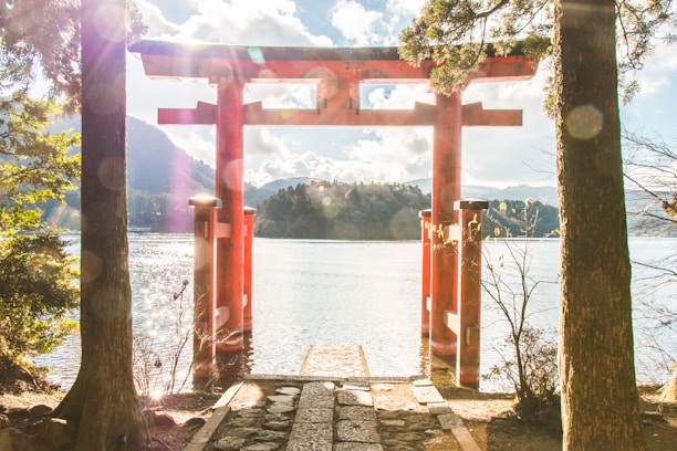 A traditional red torii gate stands prominently by the water's edge, framed by trees on either side. Sunlight filters through clouds above, creating a bright, serene atmosphere. The backdrop features a scenic view of a lake surrounded by wooded hills.