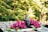 A serene setting features a dark sculpture of a face with closed eyes, surrounded by vibrant magenta flowers and lush green leaves. The sculpture is placed on a patterned surface with intricate designs, creating a calming and tranquil atmosphere. The background is filled with soft-focused greenery, adding to the peaceful ambiance.