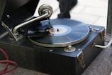A vintage gramophone with a black vinyl record spinning on its turntable. The metal tonearm and needle are positioned on the record. The wooden casing has a dark finish and is slightly worn, indicating its age.