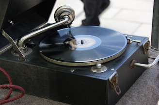 A vintage gramophone with a black vinyl record spinning on its turntable. The metal tonearm and needle are positioned on the record. The wooden casing has a dark finish and is slightly worn, indicating its age.
