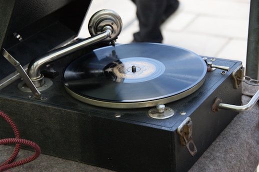 A vintage gramophone with a black vinyl record spinning on its turntable. The metal tonearm and needle are positioned on the record. The wooden casing has a dark finish and is slightly worn, indicating its age.