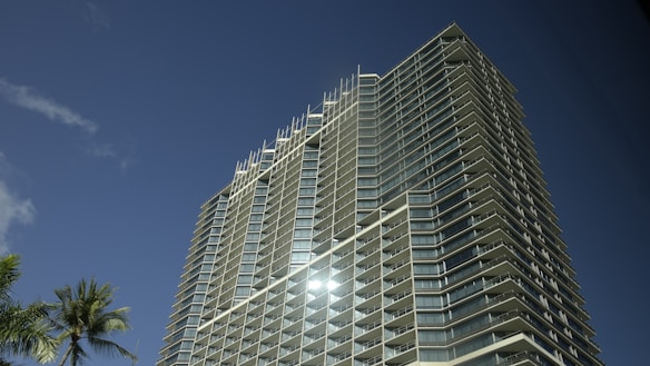 A modern high-rise building with numerous balconies is set against a clear blue sky. The sun reflects off its glass surfaces, and a palm tree is visible in the foreground.