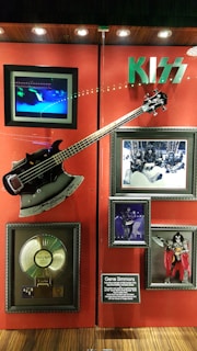 A memorabilia display featuring items related to the band Kiss. The centerpiece is a guitar resembling Gene Simmons' iconic axe, signed and mounted on a red background. Surrounding the guitar are framed photos of the band, a gold record, and a description plaque about Gene Simmons. The Kiss logo is prominently displayed above the items.