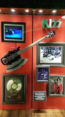 A memorabilia display featuring items related to the band Kiss. The centerpiece is a guitar resembling Gene Simmons' iconic axe, signed and mounted on a red background. Surrounding the guitar are framed photos of the band, a gold record, and a description plaque about Gene Simmons. The Kiss logo is prominently displayed above the items.