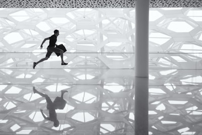 A person is mid-stride in a leap across a reflective surface with a geometric, patterned background. The reflection of the person is clearly visible on the floor, emphasizing the dynamic motion.