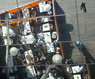 An aerial view of an outdoor dining area with several tables arranged on a wooden deck. People are seated at the tables enjoying meals, with some tables featuring shared dishes. The area is partially enclosed by an orange barrier, and the scene is reflected in glass panes with visible metal supports.