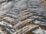 A close-up view of intricately woven ropes, displaying an interlaced pattern with varying shades of gray and hints of blue. The ropes appear to be made of a fibrous material, and the sunlight adds a subtle sheen to the texture.