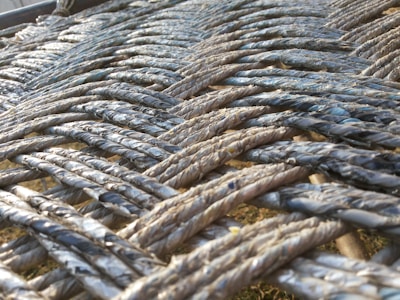 A close-up view of intricately woven ropes, displaying an interlaced pattern with varying shades of gray and hints of blue. The ropes appear to be made of a fibrous material, and the sunlight adds a subtle sheen to the texture.